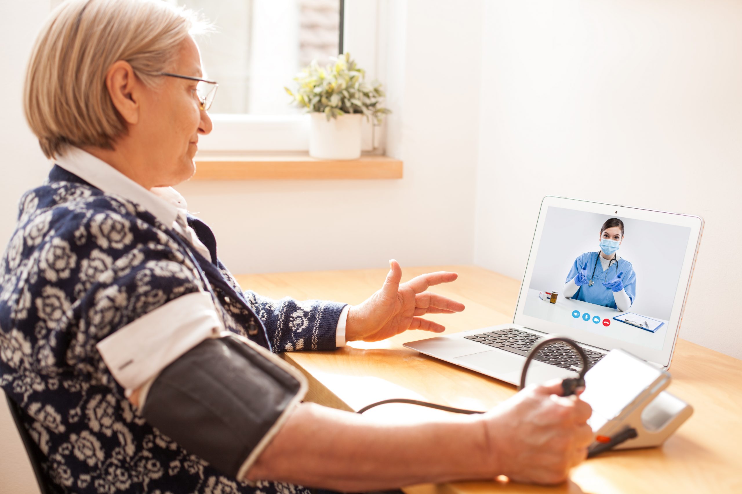 Elderly senior retired woman using sphygmomanometer blood pressure monitor to measure heart rate pulse,talking to female e-doctor via online video call help line,self-monitoring remote telemedicine